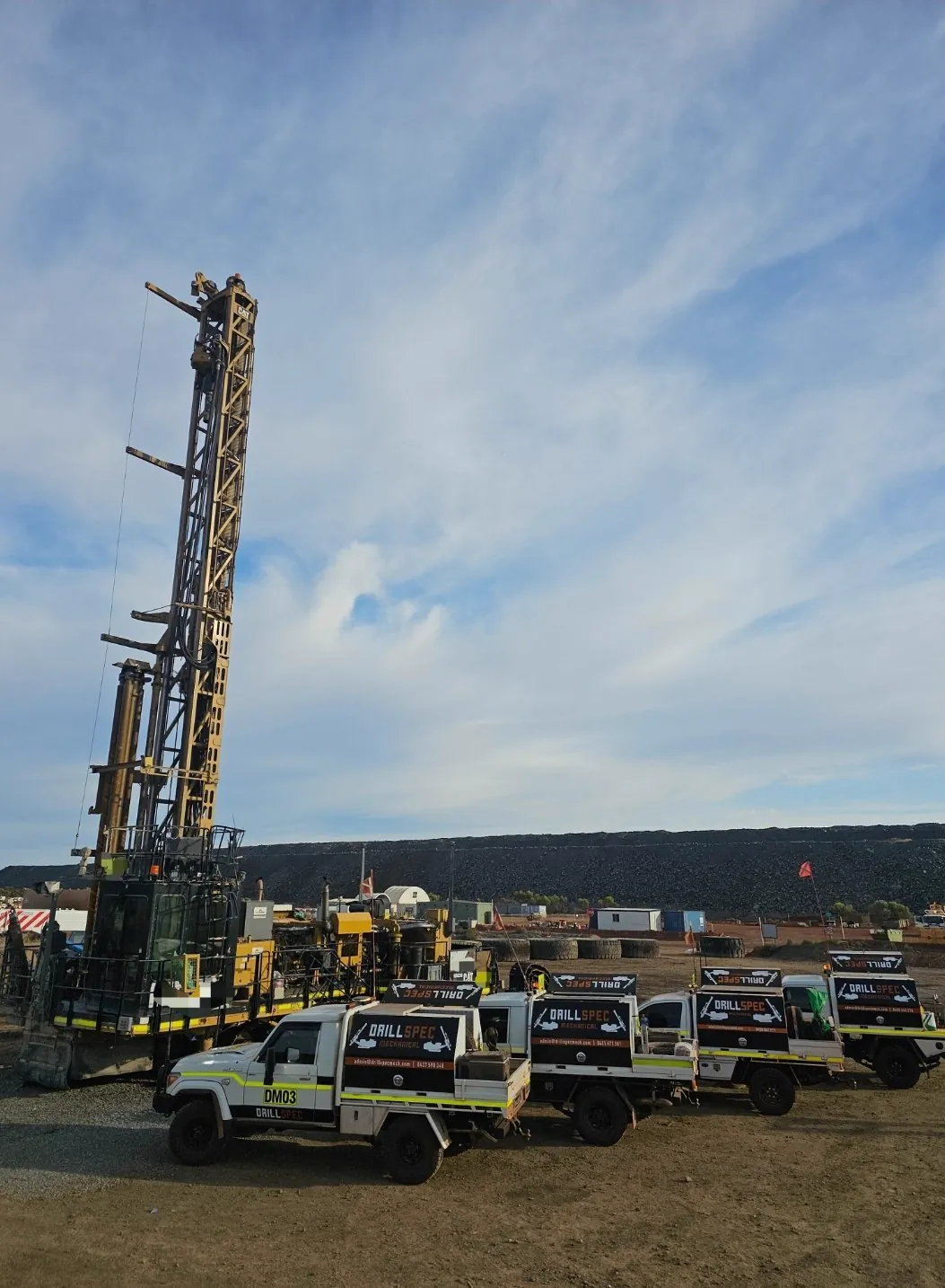 Drillspec service ute fleet (DM03 leading) lined up beside a tall CAT production drill rig at a Goldfields mine site