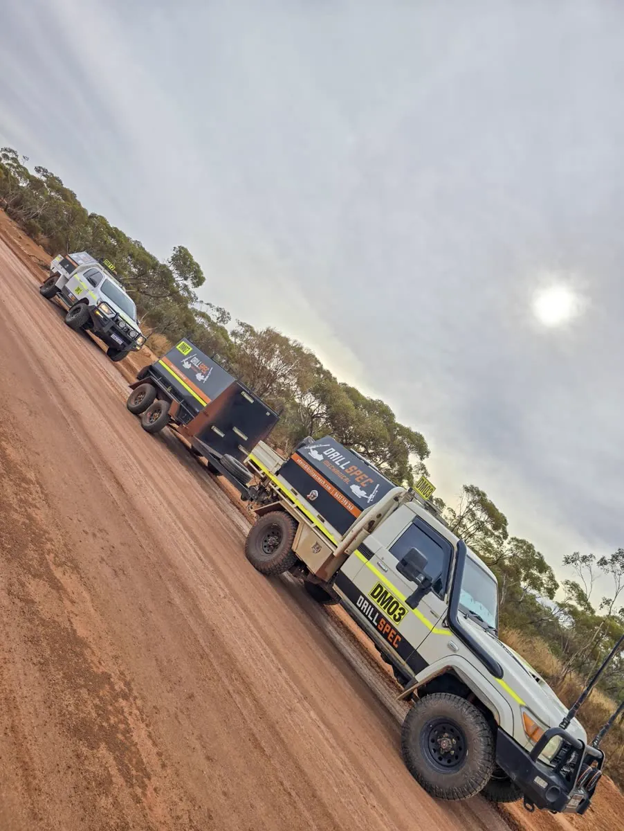 Two Drillspec service utes towing a trailer down a remote red-dirt track