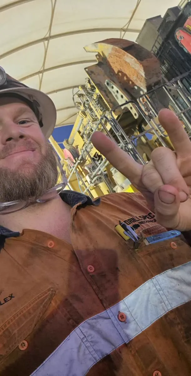 Drillspec fitter in hard hat and dirty hi-vis giving a peace sign in front of a jumbo drill under the shut dome